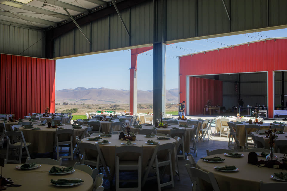 Round banquet tables set with linens and greenery inside the covered event space, open to a view of the hills.